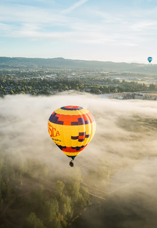 A hot air balloon soars high above the Willamette Valley. Directly below, the ground is cast with fog—but—in the distance, a quiet town and rich green hills are visible.